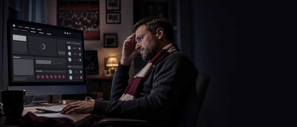 A supporter in a club scarf at a dark desk, one hand at his temple, watching a club website that has stalled on a loading spinner.